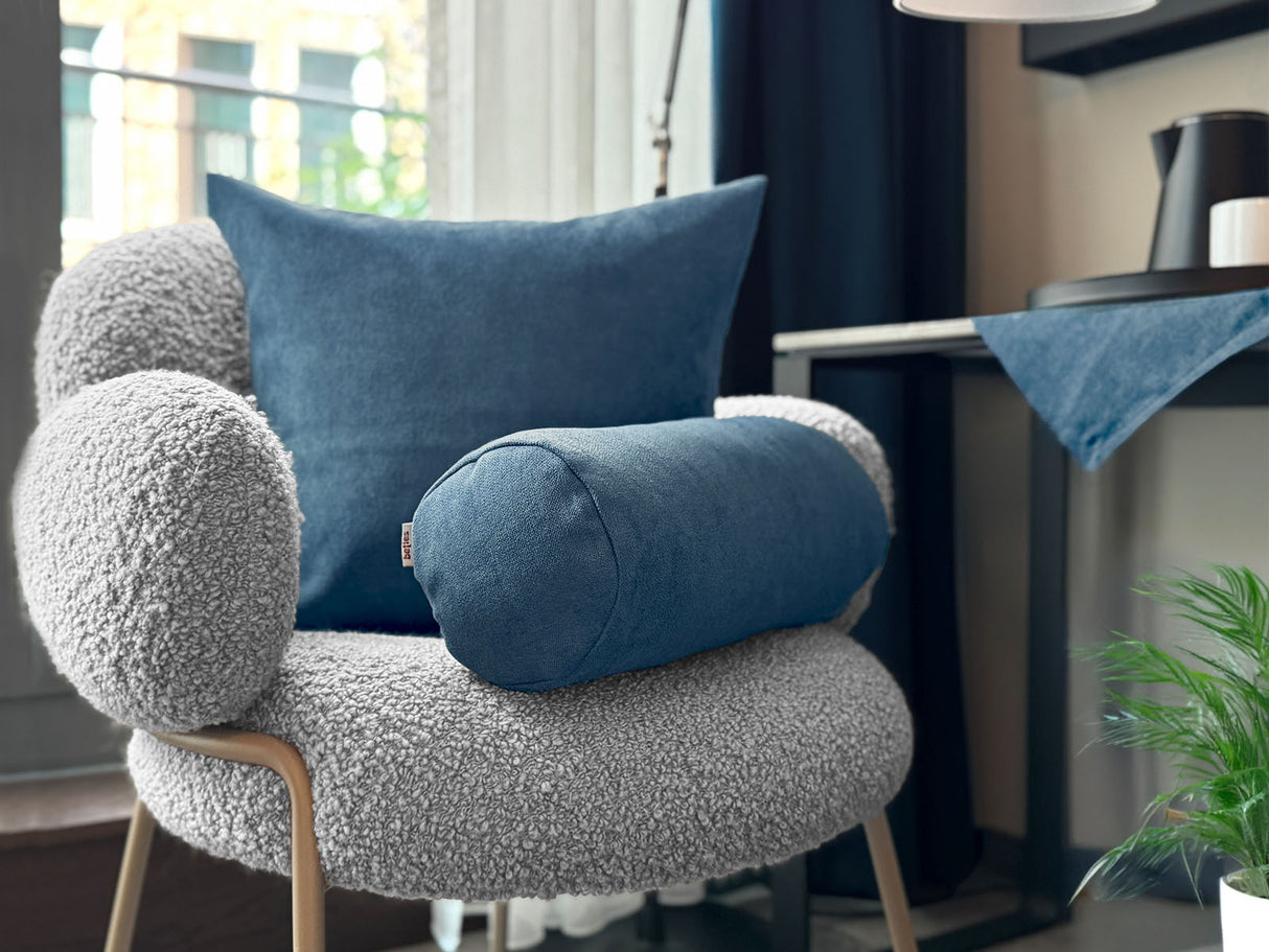 Auf einem Sessel ist ein quadratisches Kissen und eine Nackerolle in der Farbe nordlicht-blau drapiert. Im Hintergrund liegt auf einem Tisch eine Mitteldecke in der gleichen Farbe.