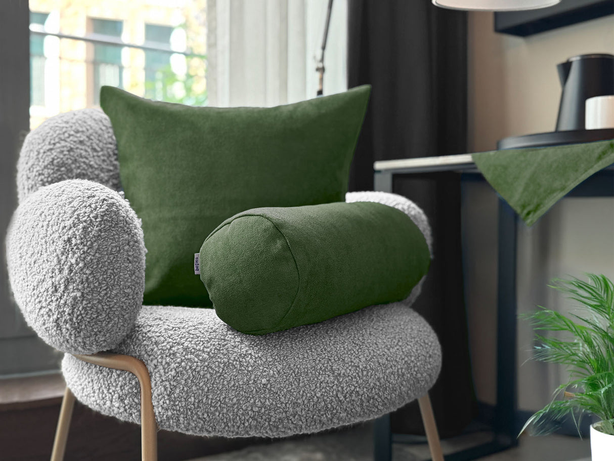 Auf einem Sessel ist ein quadratisches Kissen und eine Nackerolle in der Farbe oliven-gruen drapiert. Im Hintergrund liegt auf einem Tisch eine Mitteldecke in der gleichen Farbe.