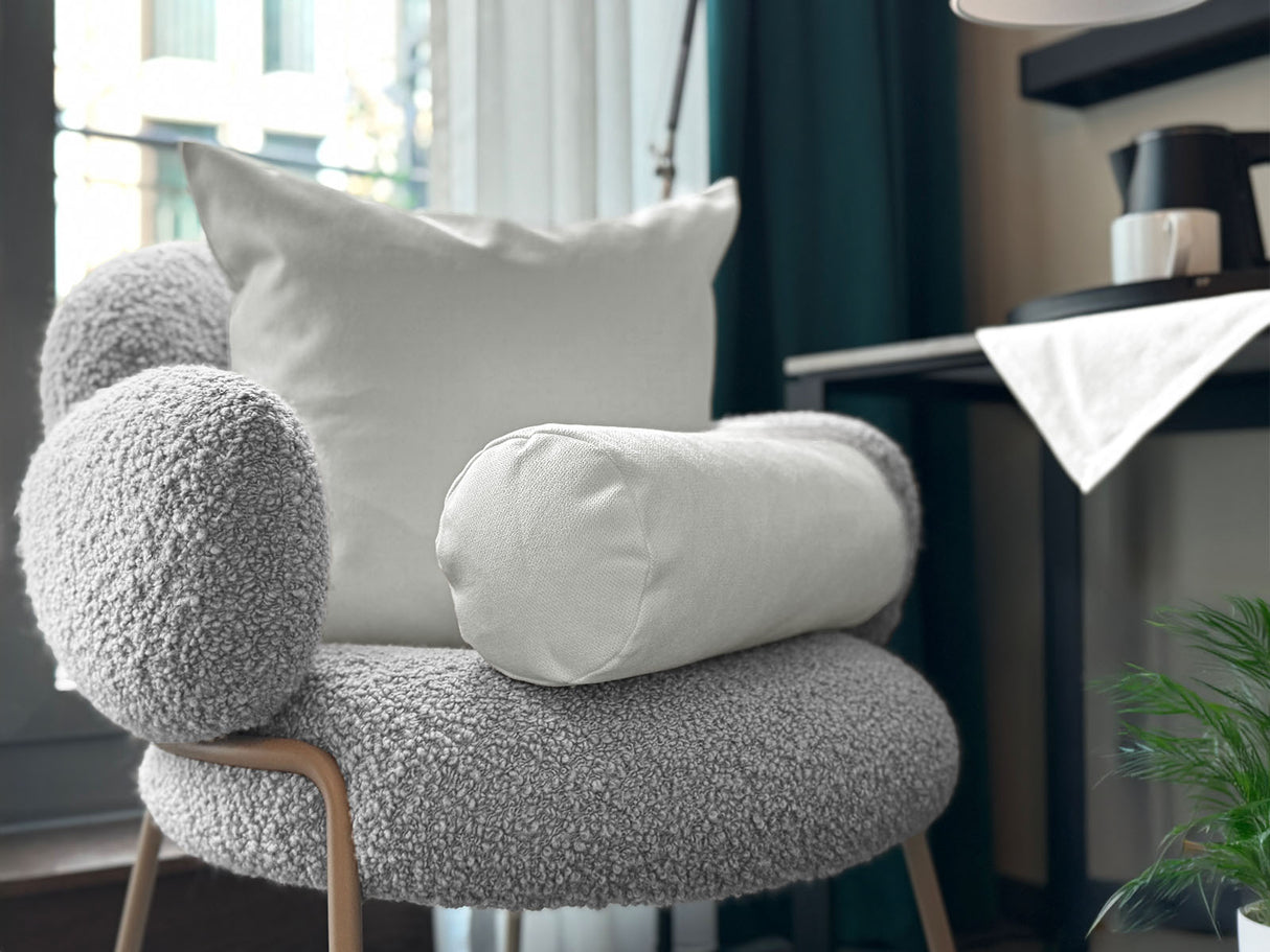 Auf einem Sessel ist ein quadratisches Kissen und eine Nackerolle in der Farbe sahne-weiss drapiert. Im Hintergrund liegt auf einem Tisch eine Mitteldecke in der gleichen Farbe.