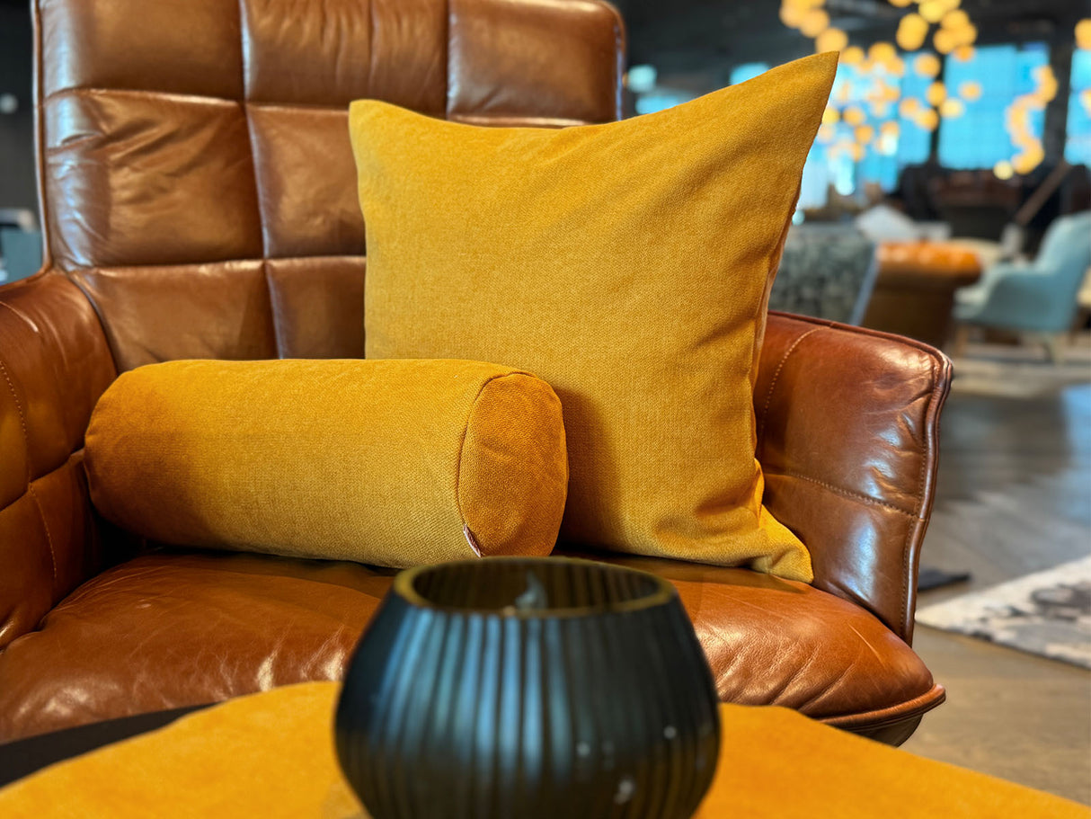 Auf einem braunen Leder-Sessel ist ein quadratisches Kissen und eine Nackerolle in der Farbe curry-gelb drapiert.