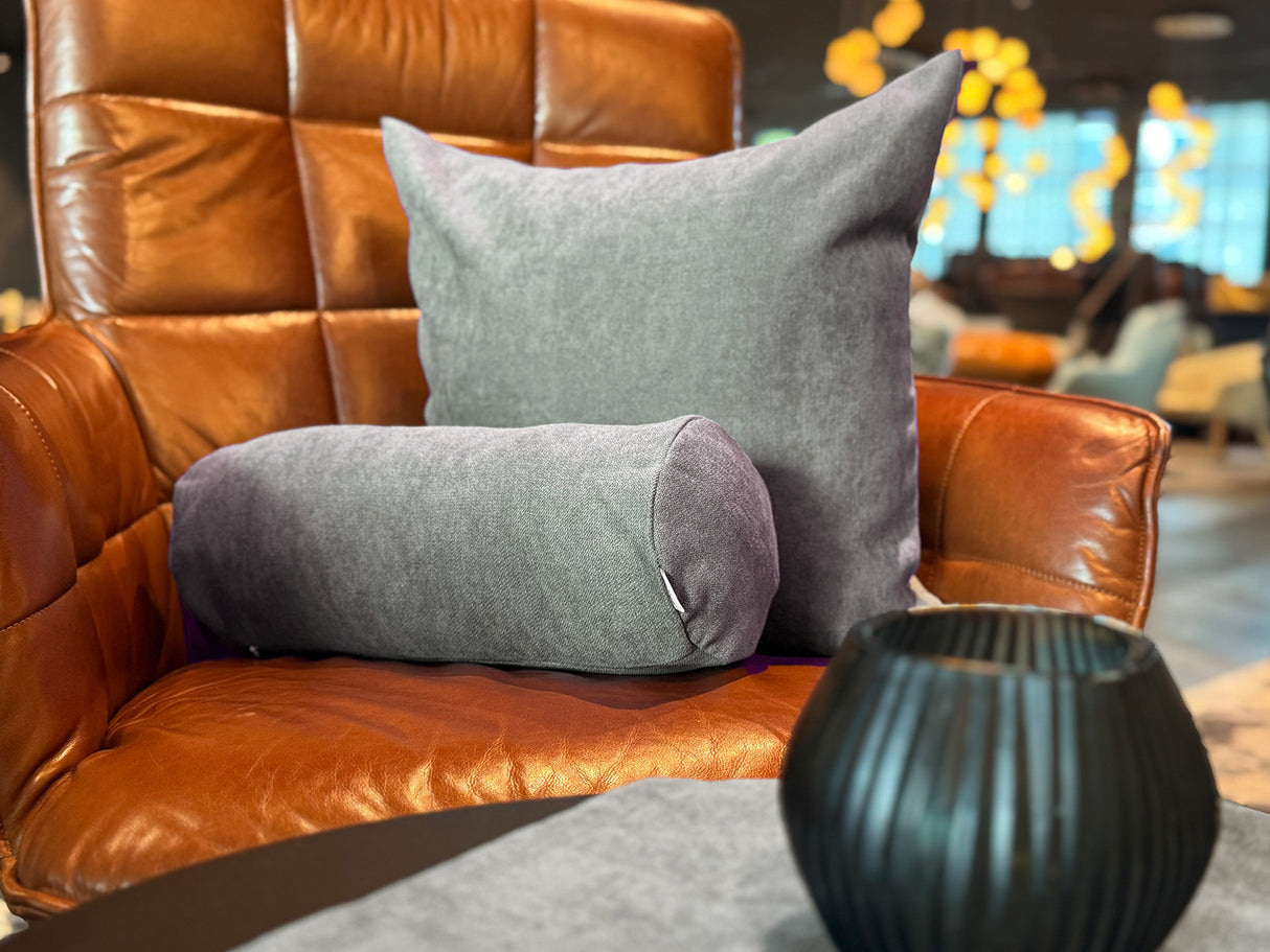 Auf einem braunen Leder-Sessel ist ein quadratisches Kissen und eine Nackenrolle in der Farbe greig-taupe drapiert.