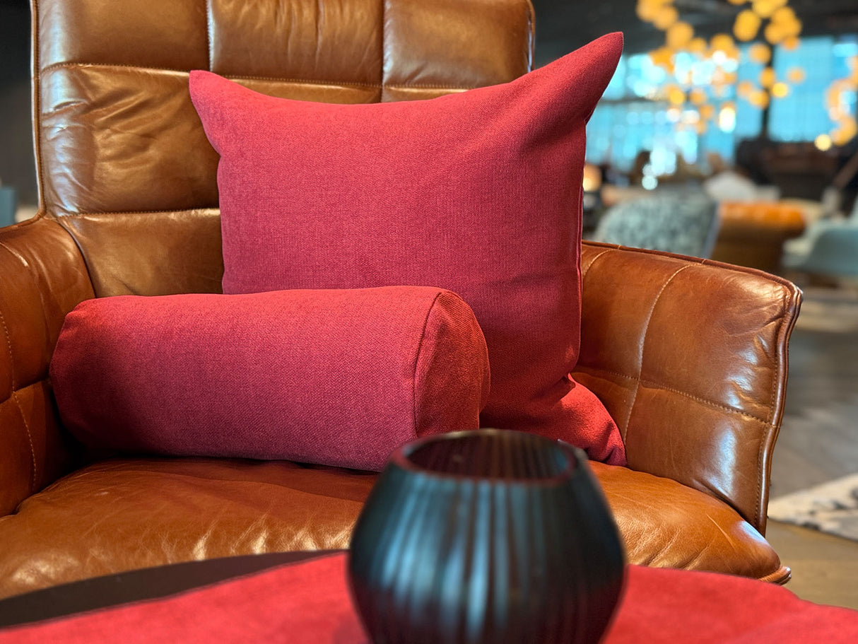 Auf einem braunen Leder-Sessel ist ein quadratisches Kissen und eine Nackenrolle in der Farbe himbeer-rot drapiert.