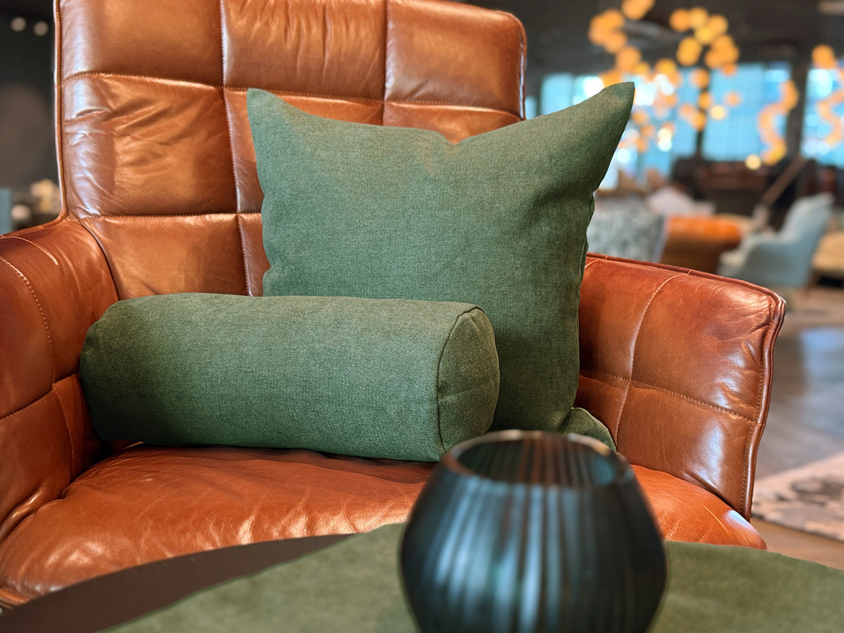 Ein quadratisches Kissen und eine Nackenrolle, drapiert auf einem Leder-Sessel, bezogen mit unifarbenen Kissenbezuegen in der Farbe oliven-gruen.