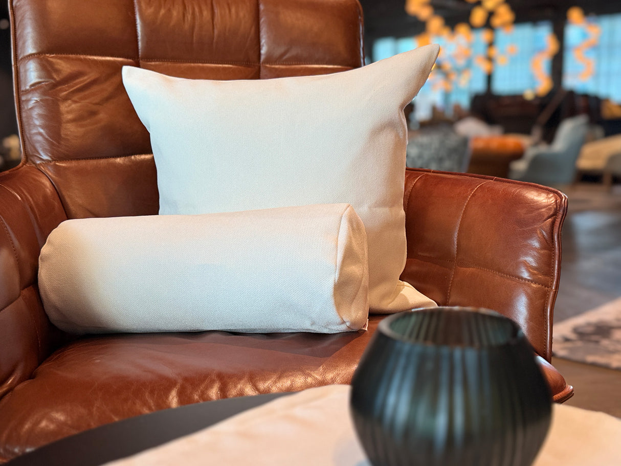 Auf einem braunen Leder-Sessel ist ein quadratisches Kissen und eine Nackenrolle in der Farbe sahne-weiß drapiert.