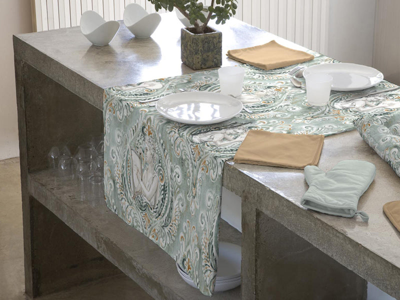 Ein gruenlicher Tischlaeufer liegt auf einem Regal. Darauf sind barocke Ornamente und griechische Goettinen mit einer Harfe abgebildet.