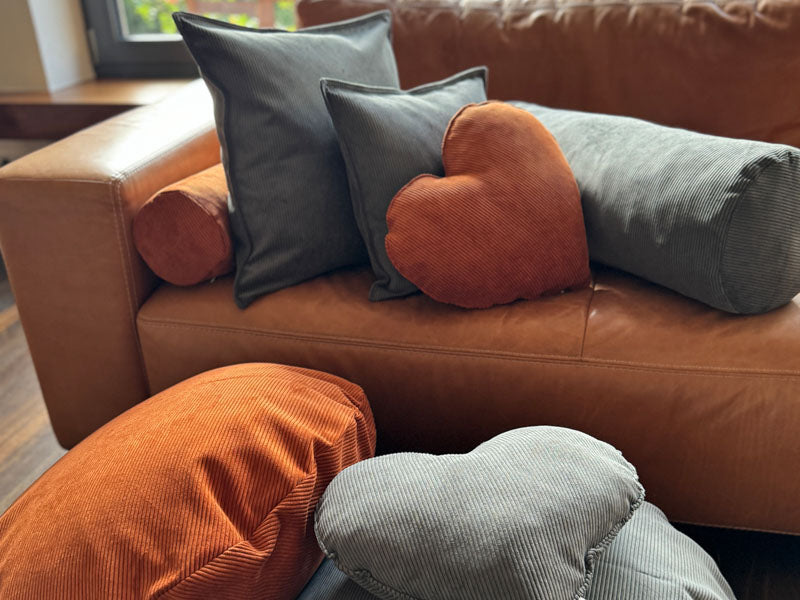 Ein braunes Ledersofa ist mit mehreren beties Hyggelig No.1 grauen und rostfarbenen Cordkissen, darunter rechteckige und herzfoermige, dekoriert, und durch ein nahe gelegenes Fenster faellt Sonnenlicht.