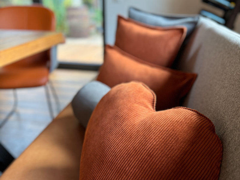 Nahaufnahme eines Sofas mit orangefarbenen und grauen beties Hyggelig No.1 Kissen, darunter ein herzfoermiges orangefarbenes Kissen, neben einem Holztisch und einem Stuhl in einem Raum mit Tageslicht.