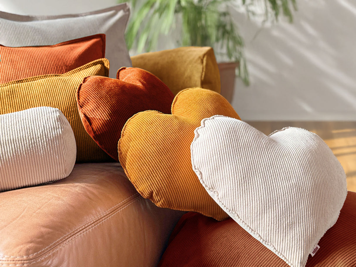 Auf einem Ledersofa liegen drei Herz-Kissen aus Cord-Samt in den Farben rost-orange, kurkuma-gelb und kreide-beige.