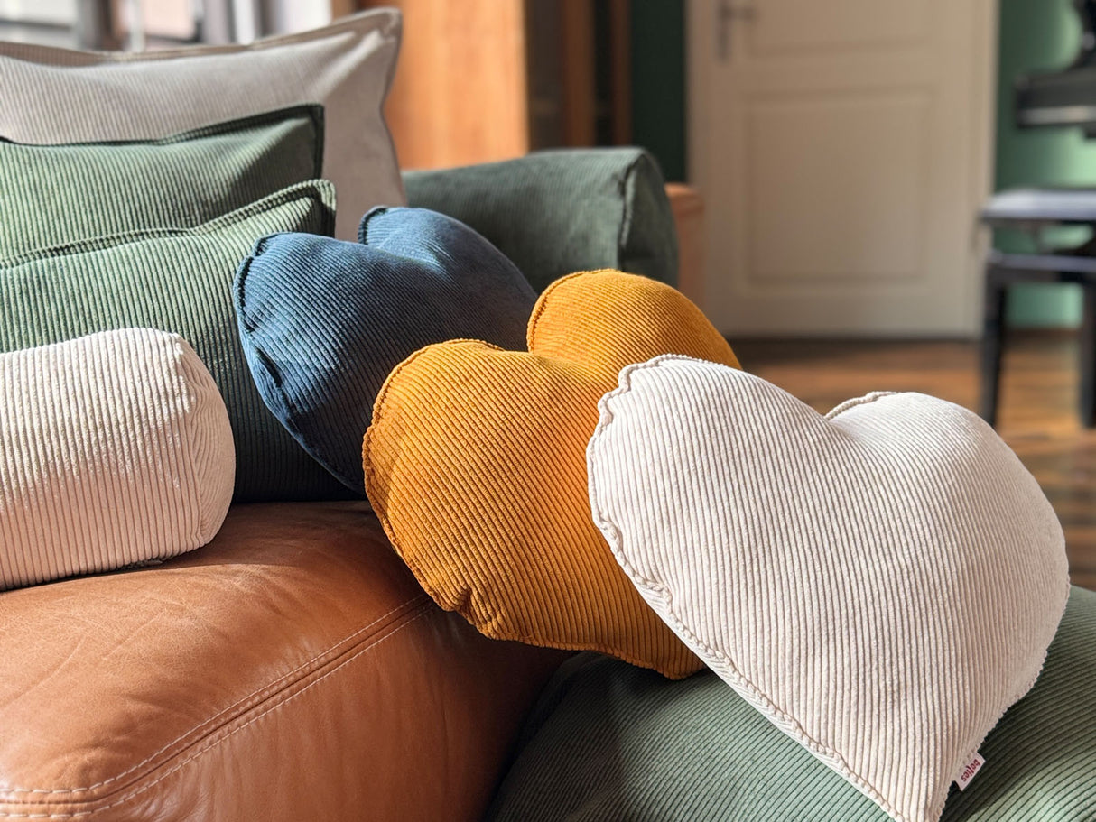 Auf einem Ledersofa liegen drei Herz-Kissen aus Cord-Samt in den Farben nord-blau, kurkuma-gelb und kreide-beige.