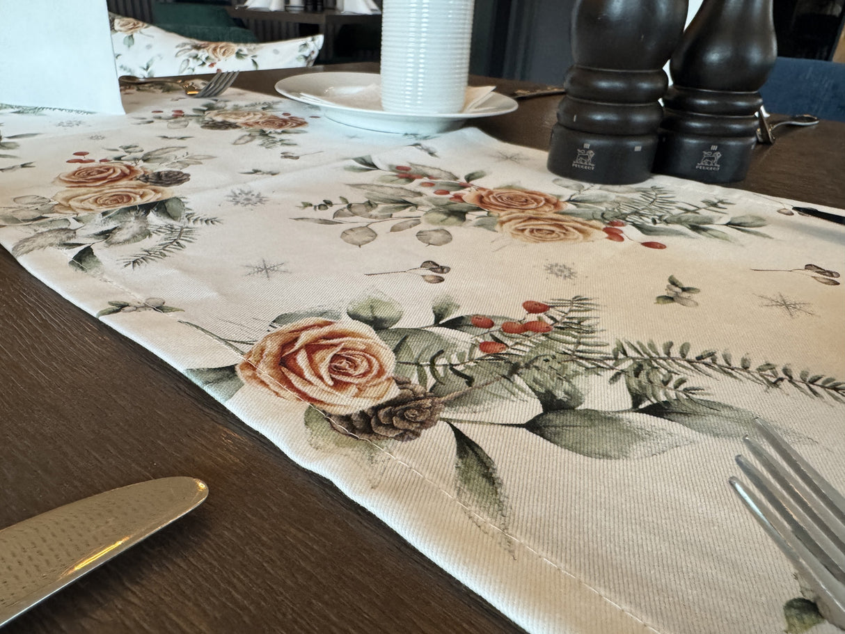 Weisser Tischlaeufer mit gefrosteten Rosen in orange, Kieferzapfen und Zweigen aus der beties Magie des Winters Kollektion "Frost-Rose" auf einem gedeckten Holztisch.