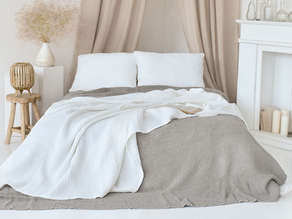 In einem Schlafzimmer liegt eine Gletscher-weisse und eine Nebel-graue Tagesdecke aus der beties "PUR.NATURE" Serie auf einem Bett ausgebreitet.