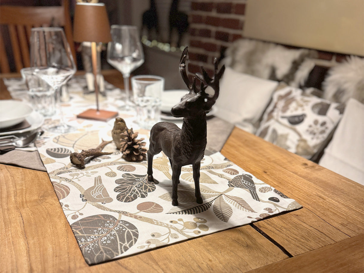 Ein eingedeckter Holztisch mit einem, mit taupefarbenen und braunen Blaettern, Zweigen und Voegeln bedrucktem, Tischlaeufer.