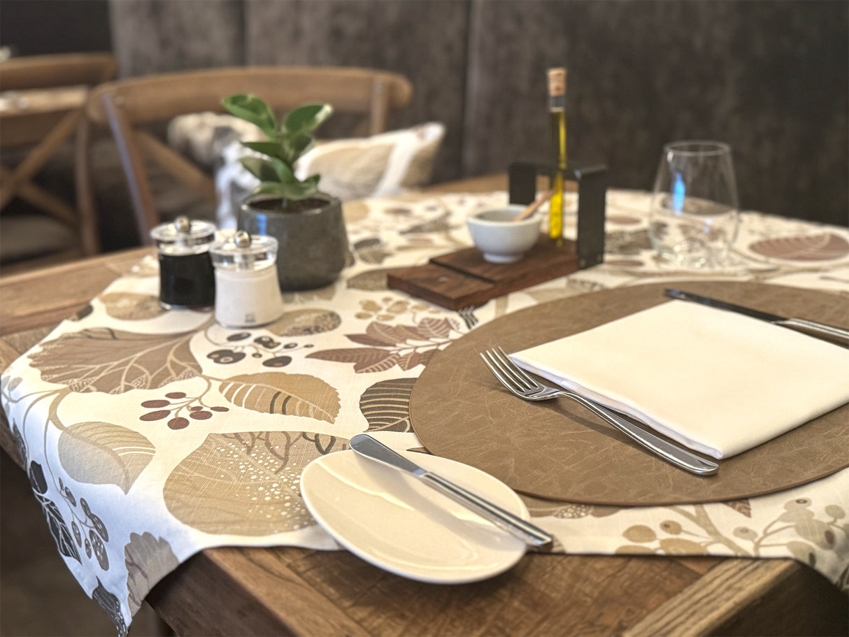 Ein eingedeckter Holztisch mit einer, mit taupefarbenen und braunen Blaettern, Zweigen und Voegeln bedruckten, Mitteldecke.