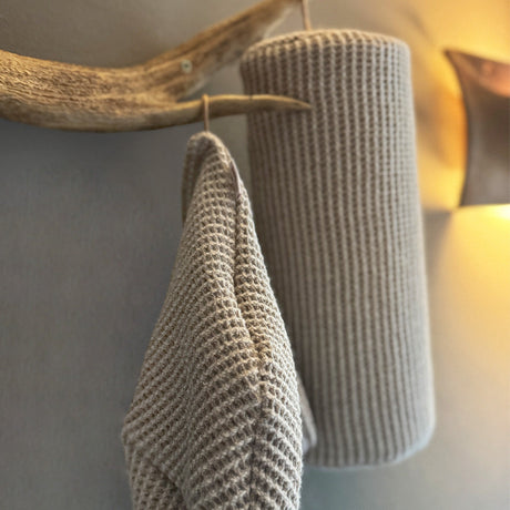 Eine Nahaufnahme von einer beigen saunsbeanie und einer Nackenrolle mit Waffelstruktur, die an einem rustikalen Holzhaken hängen, mit weicher, warmer Beleuchtung im Hintergrund.
