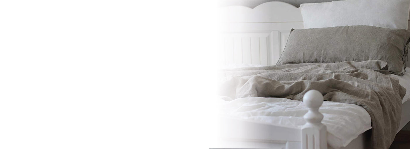 Ein fein säuberlich gemachtes Bett mit weißen Kissen und Laken, darüber eine hellgraue Leinenbettdecke mit passendem Kissenbezug, in einem sanft beleuchteten, gemütlichen Schlafzimmer. Die linke Seite des Bildes verschwindet in Weiß.
