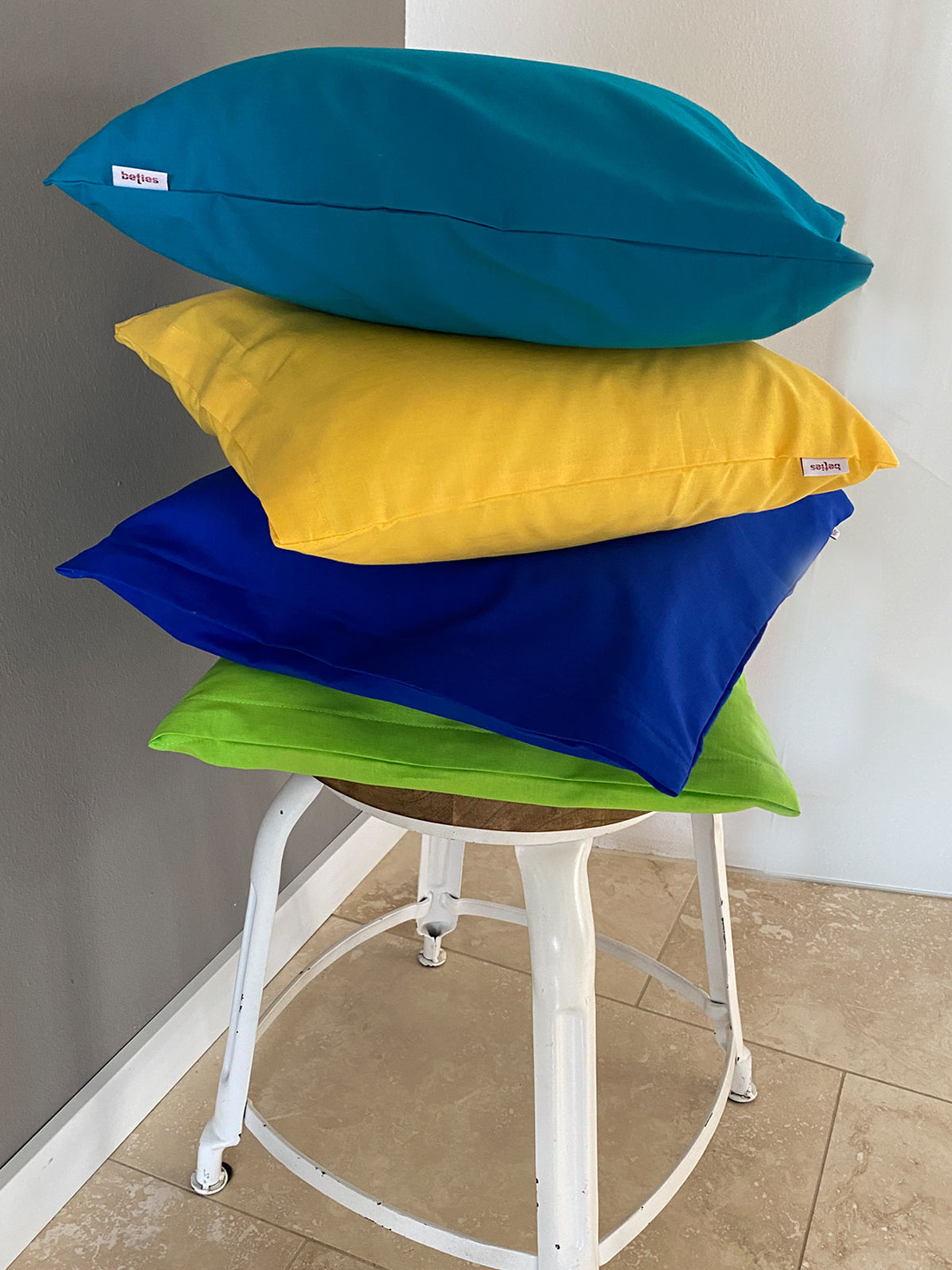 Ein Hocker mit 4 gestapelten, quadratischen Kissen mit Kissenhuellen in den Farben Gruen, Blau, Gelb und Petrol