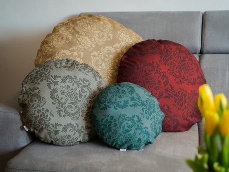 Auf einem Sofa lehnen vier runde Kissen in unterschiedlichen Groessen. Bezogen sind sie mit Bezuegen in den Farben Petrol, Grau, Rot und Beige. Bedruckt sind die Bezuege mit barocken Ornamenten in der jeweiligen Farbe, nur etwas dunkler.