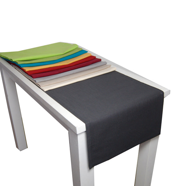 Einfarbiger, dunkelgrauer Tischlaeufer liegt auf einem weissen Tisch. Darauf liegen Farbmuster in Apfelgruen, Gruen, Petrol, Gelb, Rot, Weinrot, Grau, Beige und Hellbeige.