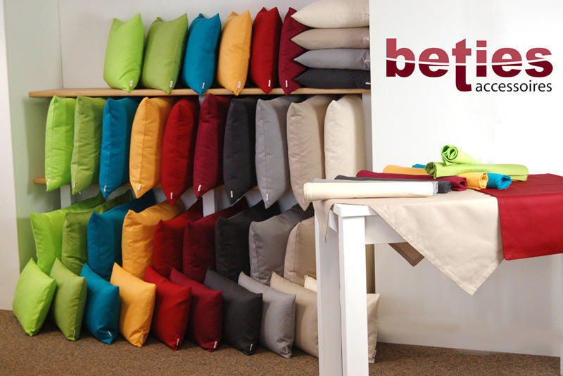 In einem Regal gestapelte, quadratische und rechteckige Kissen. Bezogen mit Kissenbezuegen in den Uni-Farben Apfel-Gruen, Gruen, Petrol, Gelb, Karmin-Rot, Hibiskus-Beere, Schiefer-Grau, Hellgrau, Beige und Hellbeige.