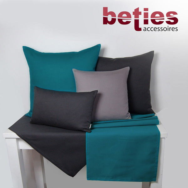 Hellbeige und Weinrote Tischwaesche auf einem weissen Tisch. Darauf stehen vier Kissen mit einfarbigen Kissenbezuegen in den Farben Hellbeige, Dunkelgrau und Weinrot.