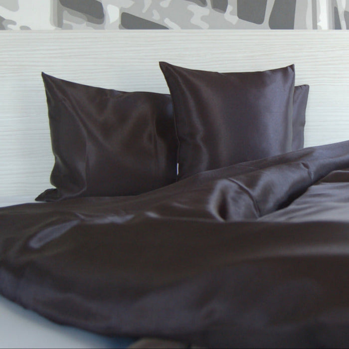 Zwei schwarze Satinkissen und ein passender Bettbezug auf einem ordentlich gemachten Bett mit einem hellen Kopfteil und einer gemusterten Wand im Hintergrund.