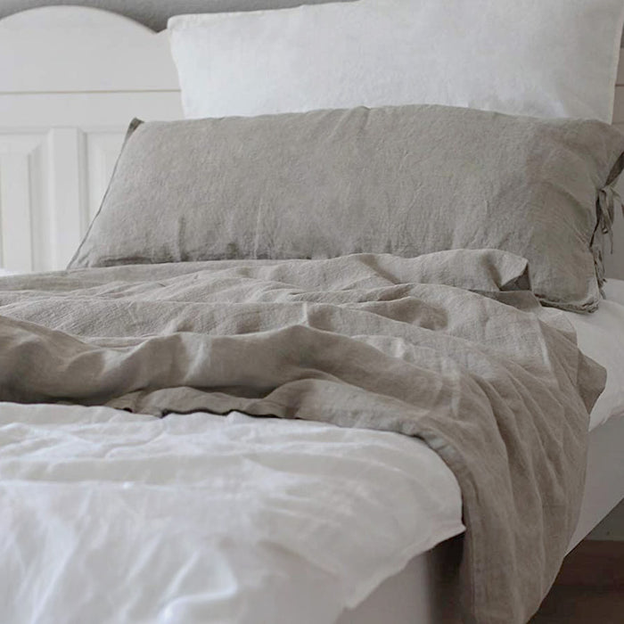 Ein fein säuberlich gemachtes Bett mit weißen Kissen und Laken, darüber eine hellgraue Leinenbettdecke mit passendem Kissenbezug, in einem sanft beleuchteten, gemütlichen Schlafzimmer.