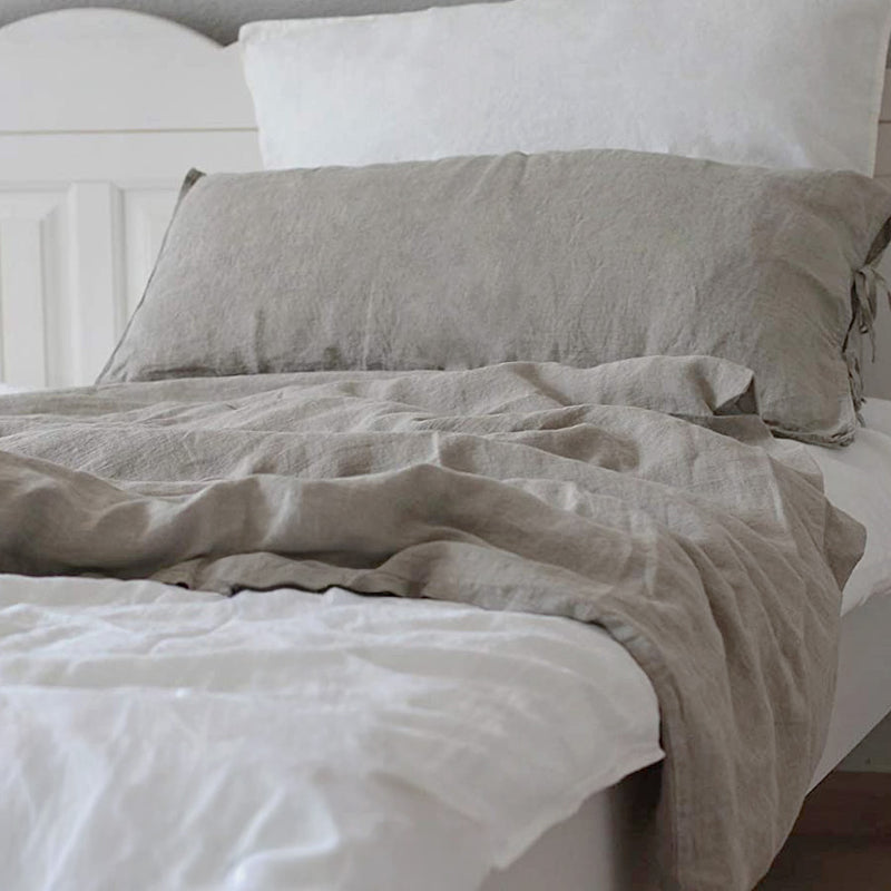 Ein fein säuberlich gemachtes Bett mit weißen Kissen und Laken, darüber eine hellgraue Leinenbettdecke mit passendem Kissenbezug, in einem sanft beleuchteten, gemütlichen Schlafzimmer. 