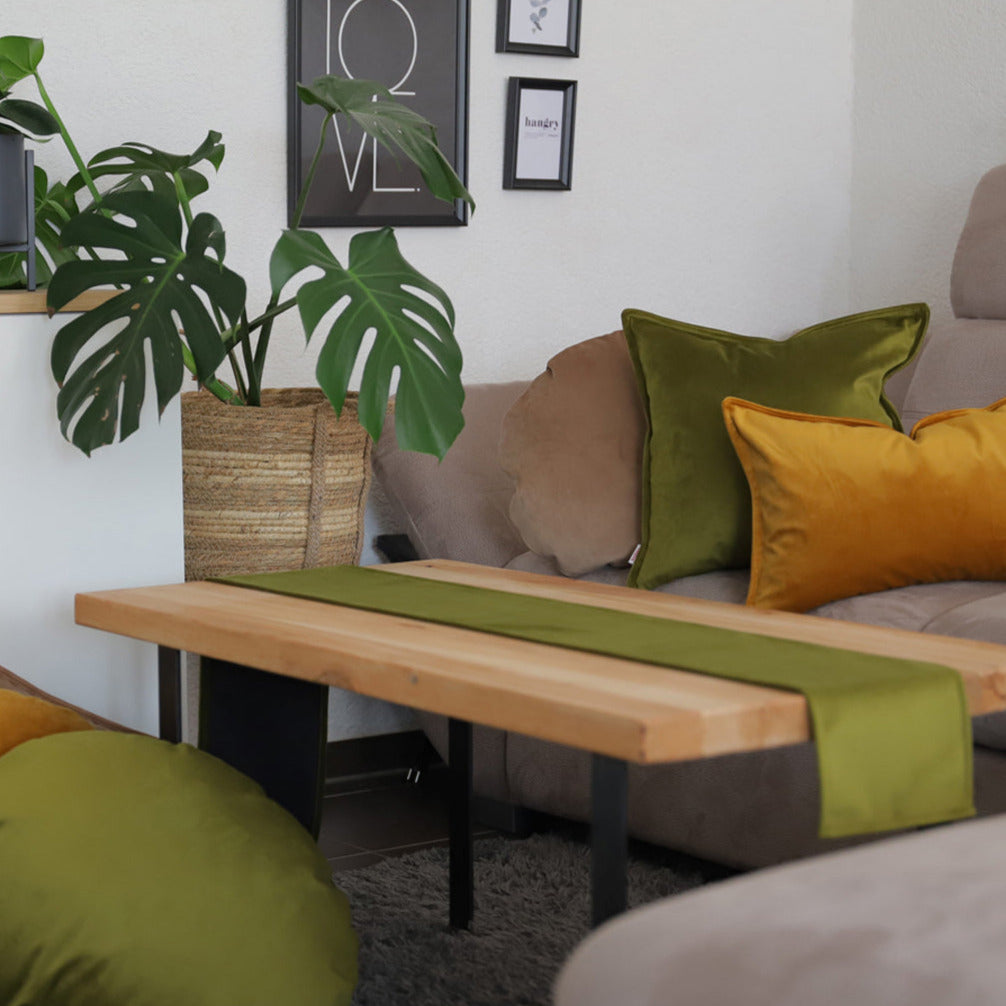 Ein gemütliches Wohnzimmer mit einem hölzernen Couchtisch, grünen und gelben Kissen, einem grünen Tischläufer und einer großen Topfpflanze neben einem beigen Sofa.