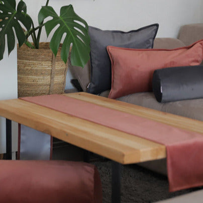Ein gemütliches Wohnzimmer mit einem hölzernen Couchtisch, grauen und rostrosanen Kissen, einem rosa Tischläufer und einer großen Topfpflanze neben einem beigen Sofa.