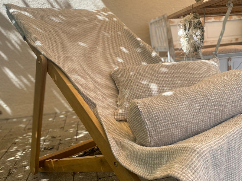 Eine Sonnenliege, die mit einem Bezug aus beigem Waffelpique bezogen ist. Darauf liegen eine Nackenrolle und ein Kissen, ebenfalls mit einem Bezug aus beigem Waffelpique-Stoff.