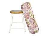 An einem Hocker lehnt ein XL-Rollen-Kissen bezogen mit einem Bezug im Landhausstil, bedruckt mit rosa/pinken Hortensien-Blueten.