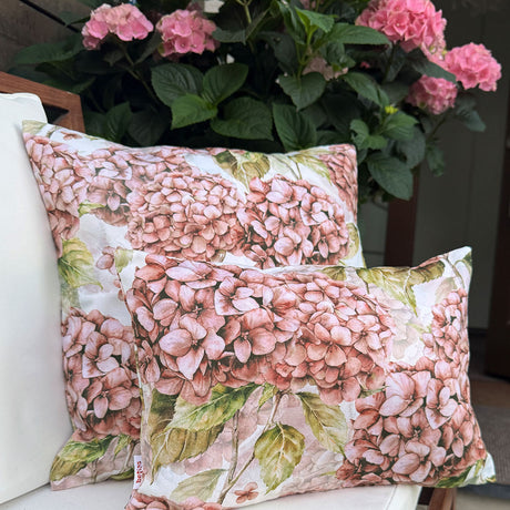 Zwei dekorative Kissen mit rosa Hortensienblumenmustern sind auf einen Stuhl gelegt, mit einer rosa Hortensienpflanze im Hintergrund.