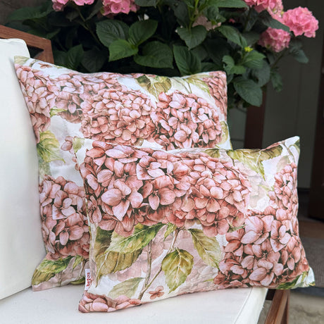 Zwei Kissen bezogen mit je einem Kissenbezug im Landhausstil, bedruckt mit rosa/pinken Hortensien. Im Hintergrund steht eine rosafarbene Hortensienpflanze.
