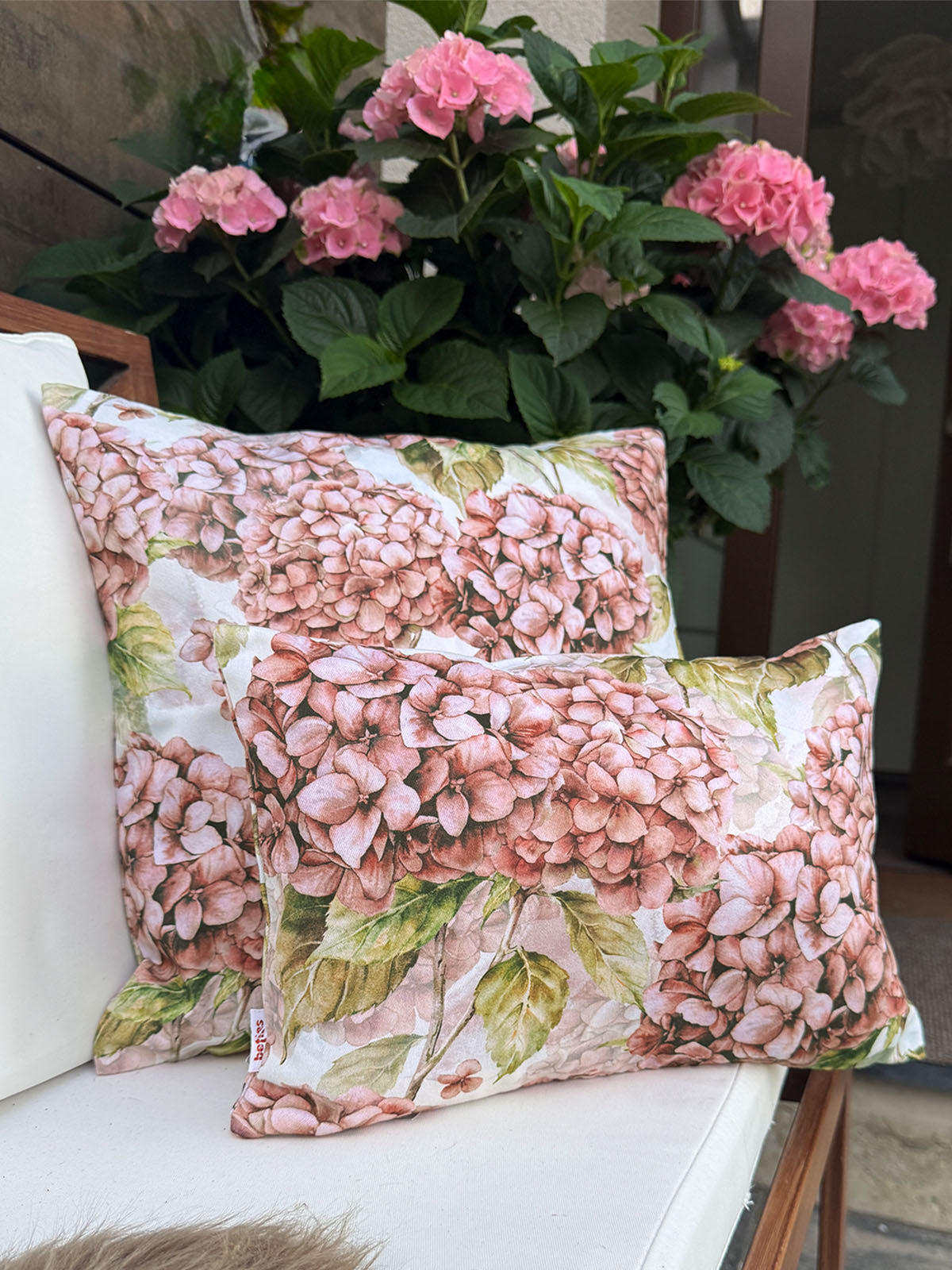 Zwei Kissen bezogen mit je einem Kissenbezug im Landhausstil, bedruckt mit rosa/pinken Hortensien. Im Hintergrund steht eine rosafarbene Hortensienpflanze.