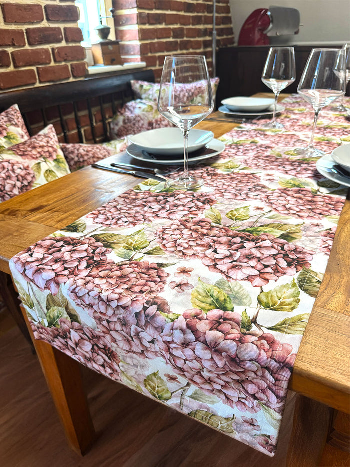 Ein hoelzerner Esstisch mit Tellern, Besteck und Weinglaesern, mit einem blumengeschmueckten Tischlaeufer und passenden Kissen mit rosa Hortensien  und gruenem Blattmustern.