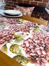 Nahaufnahme eines mit rosa/pinken Hortensien bedruckten Tischlaeufers auf einem Holztisch.