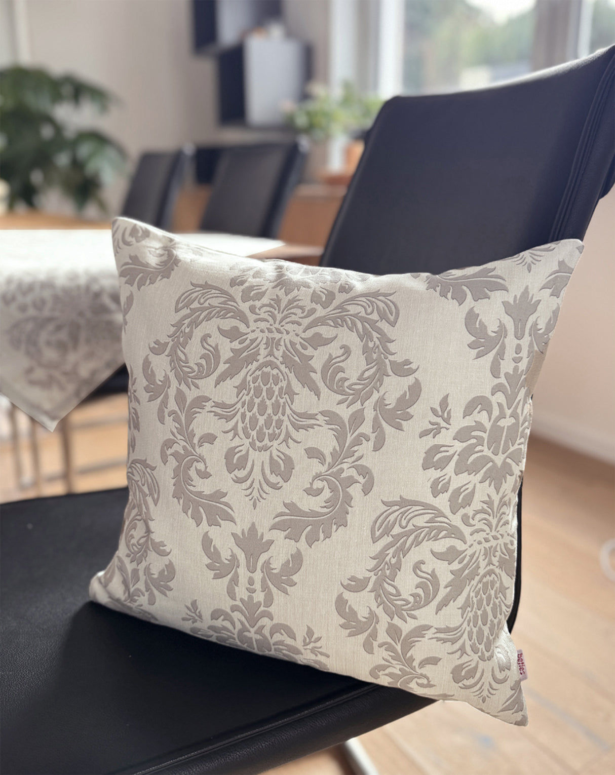 Auf einem schwarzen Lederstuhl, lehnt ein Kissen, bezogen mit einem beigen Kissenbezug der mit barocken Ornamenten in einem dunkleren Beige, bedruckt ist.