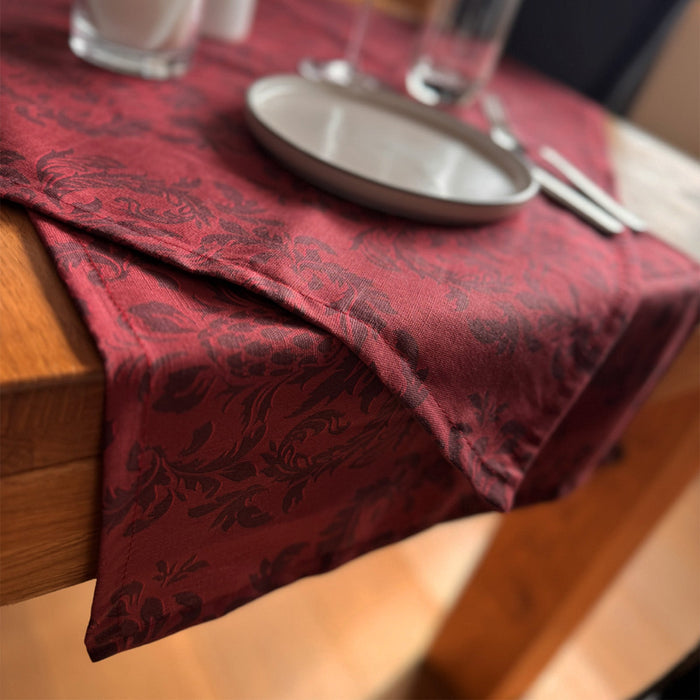Nahaufnahme eines Esstisches mit einer rot gemusterten Tischdecke, einem weißen Teller, Silberbesteck und einem Glas, alles auf einem Holztisch. Der Rand des Tischtuchs hängt über die Seite.