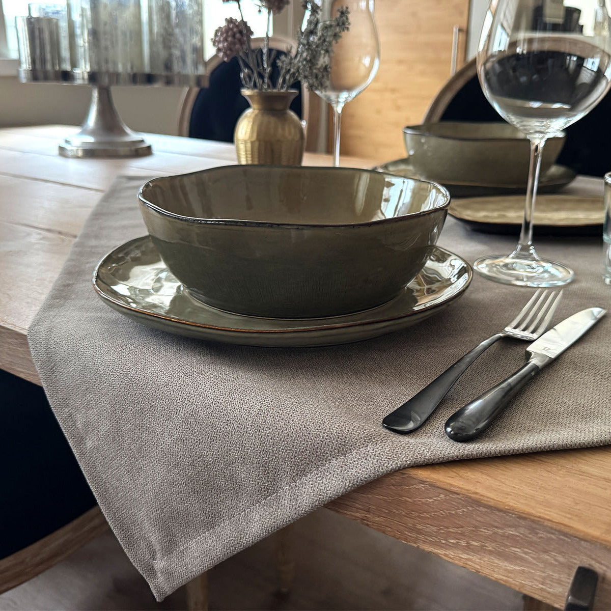 Nahaufnahme eines gedeckten Esstisches mit grauer Tischdecke, grüner Keramikschale und -teller, Silberbesteck und einem Weinglas.