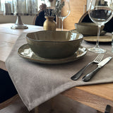 Nahaufnahme eines gedeckten Esstisches mit grauer Tischdecke, grüner Keramikschale und -teller, Silberbesteck und einem Weinglas.