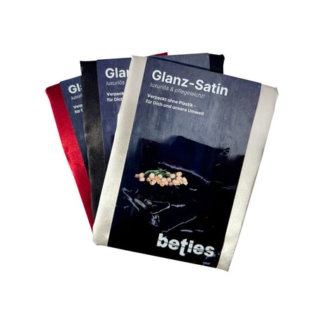 Drei gefaltete Glanz-Satin-Stoffpakete in roter, schwarzer und champagner Farbe, alle mit einem dunklen Etikett mit gedrucktem Text in deutscher Sprache, auf weißem Hintergrund