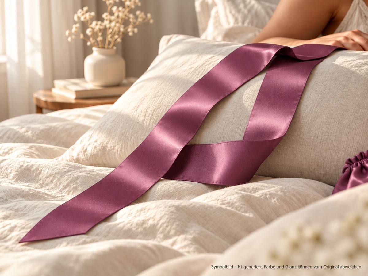 Satin-Schlafmaske in mauve locker auf einem Kopfkissen im hellen Schlafzimmer, daneben das passende Säckchen – Symbolbild, KI-generiert. Farbe und Glanz können vom Original abweichen.