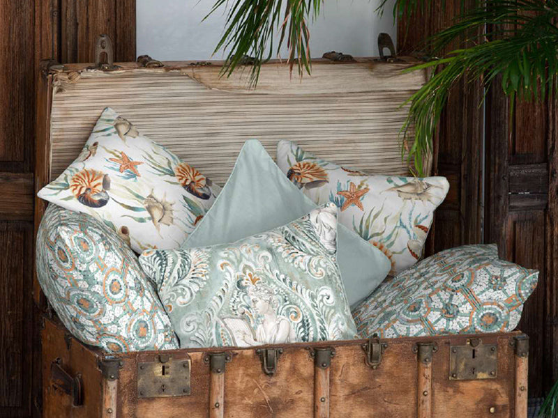 Alte Truhe gef�llt mit quadratischen Kissen. Die Kissen sind mit verschiedenen Kissenbezuegen bezogen. Auf einem ist eine G�ttin mit Harfe abgebildet. Zwei weitere sind mit Seesternen und Seeschnecken bedruckt. Andere haben Mosaik-Elemente als Muster.