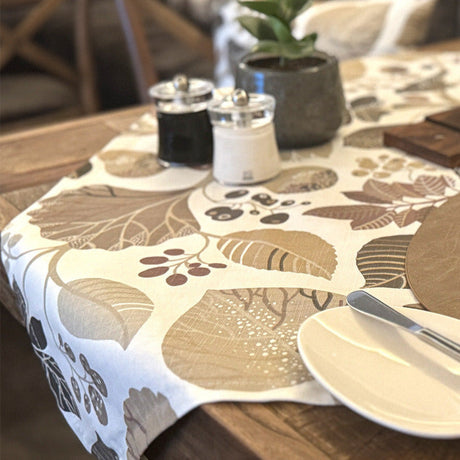 Ein hölzerner Esstisch mit einer gemusterten Tischdecke, einem Teller mit Messer und Gabel im Hintergrund. Die Tischdecke ist mit beigen und braunen Blättern gemustert.