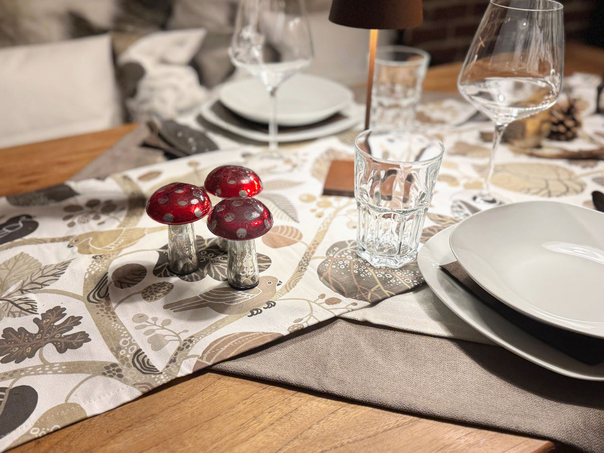 Ein eingedeckter Holztisch mit einem, mit taupefarbenen und braunen Blaettern, Zweigen und Voegeln bedrucktem, Tischlaeufer.